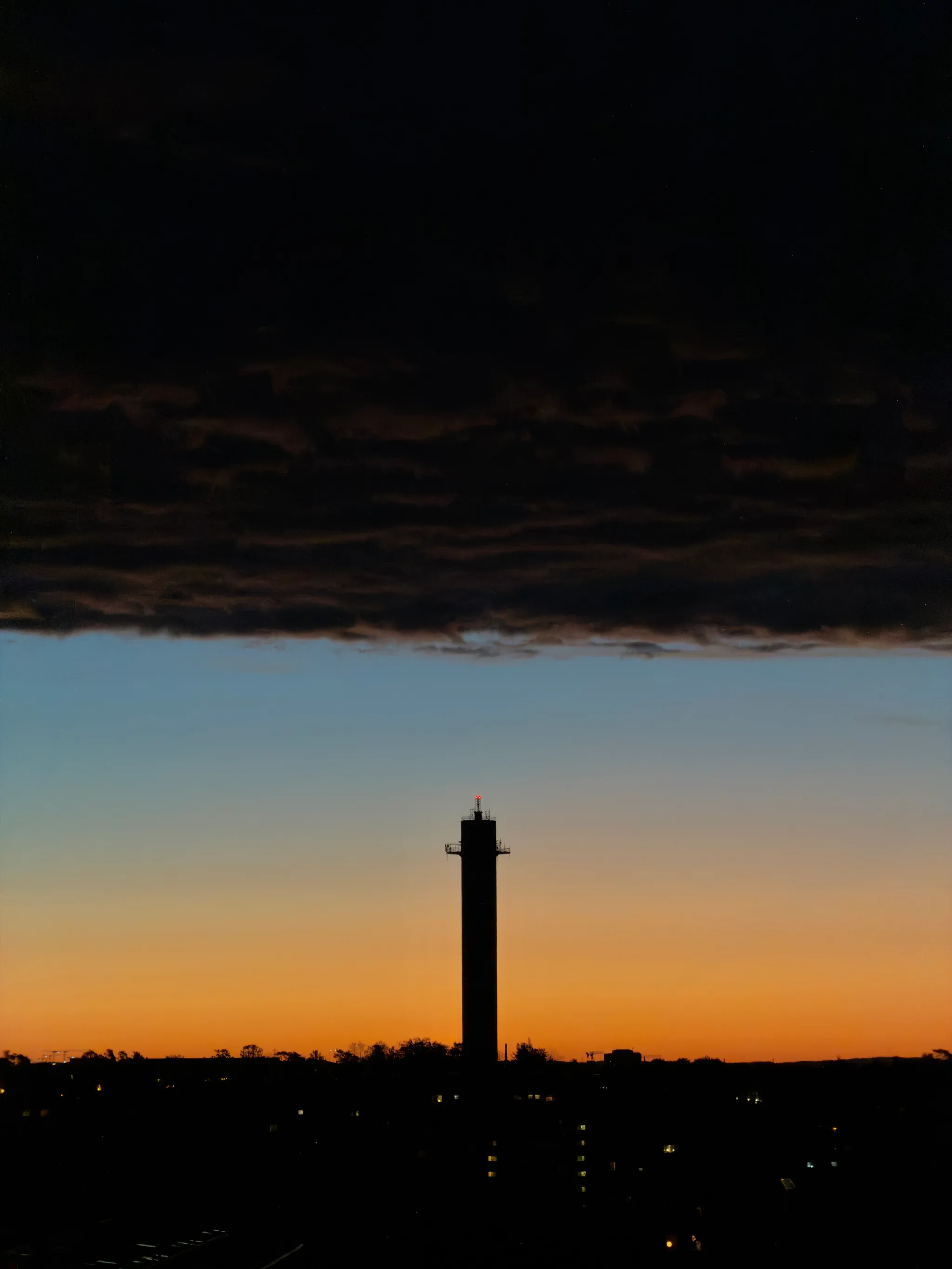 A slender tower silhouetted with a red beacon light rises against a glowing orange and blue dusk sky, beneath heavy dark clouds, with scattered lights along the horizon.